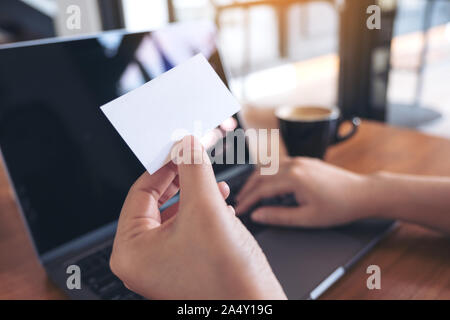 Eine Hand, die eine weiße Leere Visitenkarte bei der Verwendung von Laptops mit Kaffee Tasse auf hölzernen Tisch in Office Stockfoto