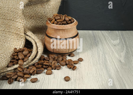 Kaffeebohnen überlaufen von einem kleinen hölzernen Topf neben einem Jute Sack in einem rustikalen noch Leben auf einem Holztisch mit Platz kopieren Stockfoto