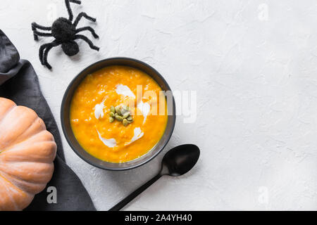 Halloween Kürbis und Karotten Suppe mit Sahne in schwarz Schüssel eingerichtet Spielzeug Spinne auf grauem Stein Hintergrund. Ansicht von oben und Platz für Text. Stockfoto