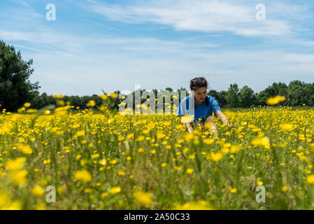 Low Angle View der jugendlich Mädchen mit geflochtenem Haar sitzt im großen Feld mit gelben Blumen und Bäume im Hintergrund Stockfoto