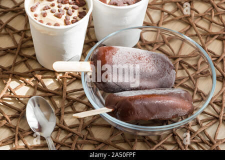 Gesund vegan hausgemachten Dessert auf Rattan Tischdecke. Raw Bananen-eis beschichtet mit natürlichen Schokolade in einem White Paper Cup und Schokolade Eis am Stiel Stockfoto