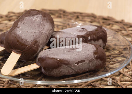 Köstliche vegane hausgemachte Desserts. Frischen, rohen Eis am Stiel auf Sticks umhüllt mit Schokolade Glasur lag in Glas transparent Schüssel auf Rattan Tischdecke. Top Stockfoto