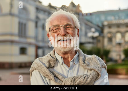 Für immer jung. Portrait von gut aussehenden Bärtigen älteren Mann in Gläsern an Kamera schauen und lächeln beim Stehen im Freien Stockfoto