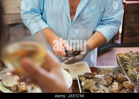 Entfernen der Schalen frische Austern mit einer Auster Messer auf einem Fischmarkt. Menschen mit einem Glas Champagner oder Sekt Stockfoto