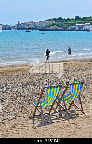 Zwei Menschen, Mann und Frau, zu Fuß in Richtung einander am Wasser am Sandstrand. Zwei leere gestreifte Liegestühle im Vordergrund. Stockfoto