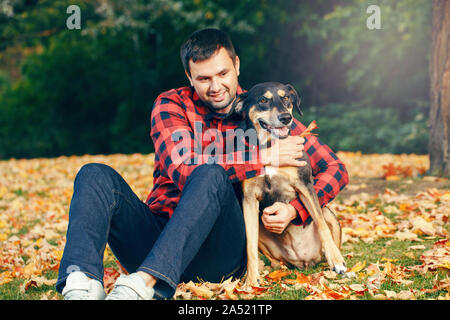 Glückliche junge Kaukasier bärtiger Mann mittleren Alters saß auf dem Boden unter den Herbst gelbe Blätter und umarmen Haustier Hund. Beste Freunde Spaß im Freien. F Stockfoto