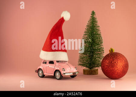 Rosa Retro Spielzeug Modell Auto mit kleinen roten Weihnachten Santa Claus hat und wenig Xmas Tree, rot Christbaumkugel auf Pastell rosa Hintergrund Stockfoto