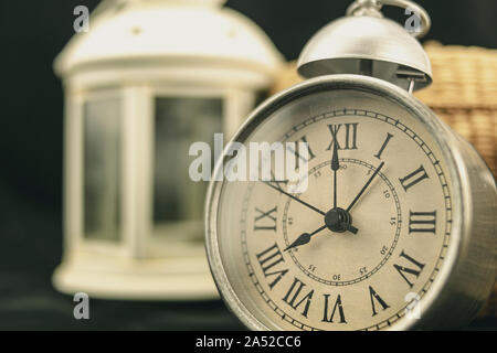 Grau vintage Uhr mit einem Weidenkorb und eine klassische weiße Laterne hinter sich, auf einem schwarzen Hintergrund Stockfoto