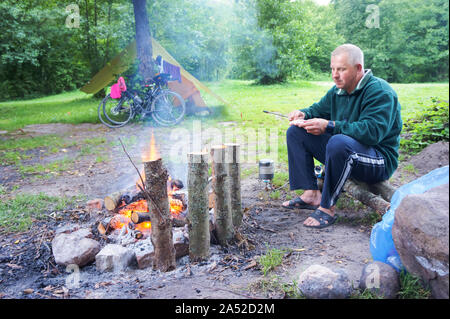 Ein Mann braten Würstchen am Lagerfeuer, Zelt, Fahrräder und Bonfire, Region Kaliningrad, Russland, 1. August 2019 Stockfoto