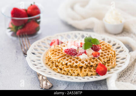 Schwedische Waffeln mit Erdbeeren und Sahne. Es ist eine Glasschale mit ganzen Erdbeeren und einem keramischen kleinen Schüssel Schlagsahne im Hintergrund Stockfoto