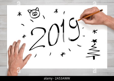 Männliche Hände halten Bleistift vor weißen Blatt Papier mit '2019'-Schild, Schwein Gesicht, Weihnachtsbaum und Schneeflocken auf gezeichnet Stockfoto