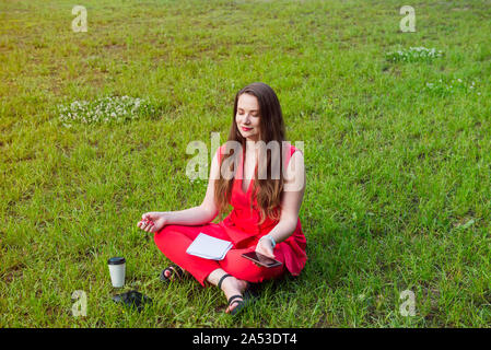 Junge Geschäftsfrau im roten Anzug sitzt im Lotussitz auf grünem Gras in Park und halten Sie Ihr Smartphone. Meditation für Lfe. Ruhige, entspannte lifest Stockfoto