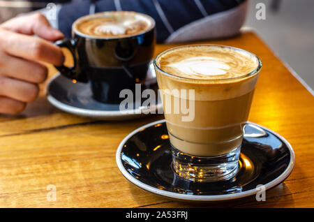 Nahaufnahme einer Tasse Café Latte mit einem verschwommenen Hintergrund der Hand eines Mannes, der seinen Kaffee auf der gegenüberliegenden Seite des Tisches hält. Stockfoto