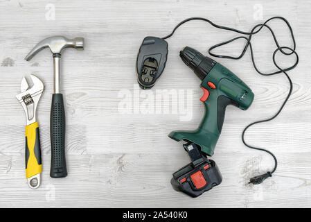 Ein Akku-bohrschrauber, es Ladegerät neben einen Hammer und einen Schraubenschlüssel an den hölzernen Tisch Hintergrund. Stockfoto