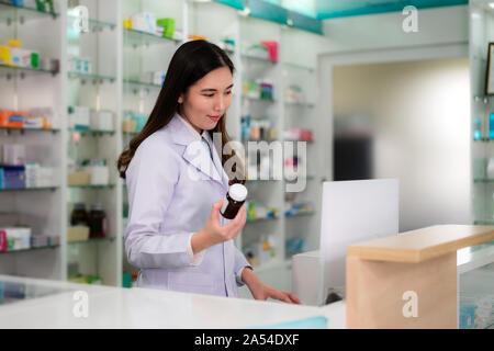 Zuversichtlich asiatische Junge weibliche Apotheker mit Smile Holding eine Medizin Flasche und Suche das Produkt auf dem Computer Datenbank in der Apotheke Drogen Stockfoto