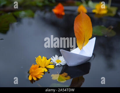 Ein Papier Boot schwimmt im dunklen Wasser, und um helle Blumen und Laub. Stockfoto