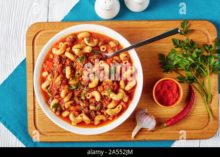 Klassische amerikanische Gulasch Ellenbogen Nudeln, Rindfleisch, Sellerie und Tomaten in einer weißen Schüssel mit Löffel, auf einer hölzernen Schneidebrett, Ansicht von oben, flatlay, Stockfoto