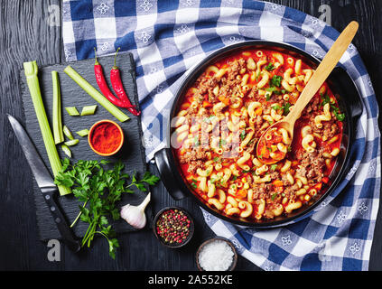 In der Nähe von amerikanischen italienischen Gulasch Ellenbogen Nudeln, Rindfleisch, Sellerie und Tomaten in einer schwarzen Keramik Teller auf einem rustikalen Holztisch mit Zutaten auf einem Stockfoto