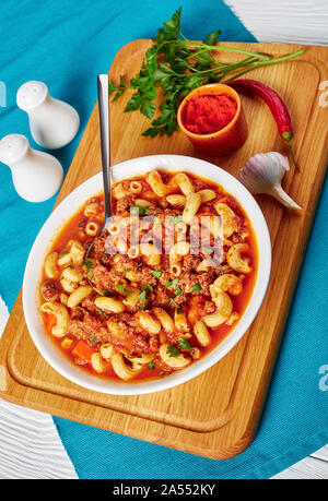 Gulasch Ellenbogen Nudeln, Rindfleisch, Sellerie und Tomaten in einer weißen Schüssel mit Löffel, auf einer hölzernen Schneidebrett, Ansicht von oben, Nahaufnahme, vertikale Ansicht Stockfoto