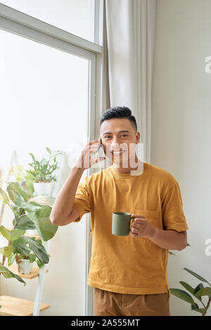 Junger Mann legere Kleidung sprechen auf ein Handy am Morgen an einem Fenster Stockfoto