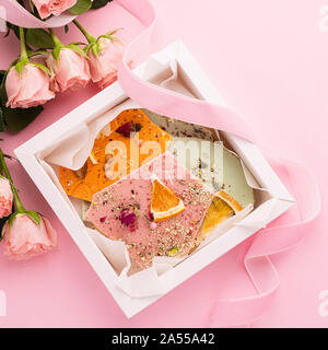 Handgemachte Schokolade Stücke in verschiedenen Farben auf einem rosa Hintergrund und ein Strauß Rosen. Exklusive dessert Geschenk. Urlaub Konzept. Quadratischen Rahmen. Stockfoto