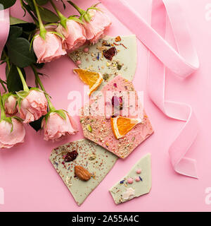 Handgemachte Schokolade Stücke in verschiedenen Farben auf einem rosa Hintergrund und ein Strauß Rosen. Exklusive dessert Geschenk. Urlaub Konzept. Quadratischen Rahmen. Stockfoto