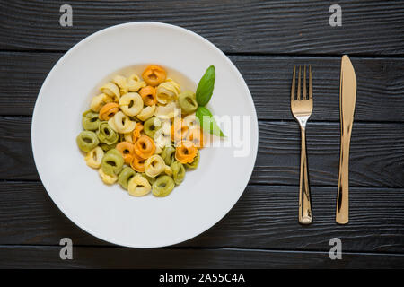 Tortellini Nudeln mit Basilikum Blatt auf serviert Tisch mit Messer und Gabel. Gesunde italienische Küche auf schwarzen Holztisch Hintergrund. Stockfoto
