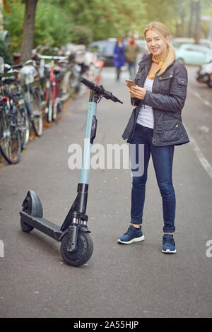 Junge blonde Frau, die in der Nähe von elektrischen kick Scooter auf Stadt Straße oder den Park, mit Fahrrädern auf der Seite geparkt. Telefon in den Händen halten und Lächeln Stockfoto