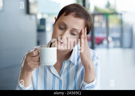 Business woman Gefühl von der Arbeit im Büro betont. Frauen mittleren Alters mit Kopfschmerzen und trinken Kaffee oder Tee Stockfoto