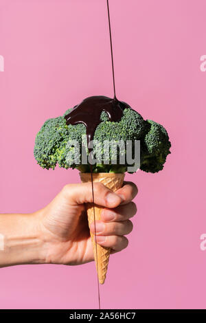 Studio shot der Hand Mann hält Eistüte mit Brokkoli auf und gießen Schokoladensauce, gegen rosa Hintergrund Stockfoto