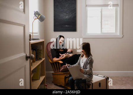 Junge Frau mit Baby Sohn im Wohnzimmer Sessel, während Tochter Buch liest Stockfoto