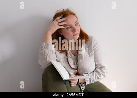 Junge Frau mit roten Haaren sitzen Holding Tagebuch und weg schauen zu Hause Stockfoto