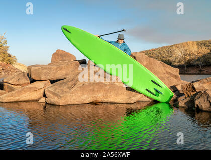 Ältere männliche Paddler stößt eine aufblasbare Stand up paddleboard von einem felsigen Ufer des Bergsee - horsetooth Reservoir in Northern Colorado Stockfoto