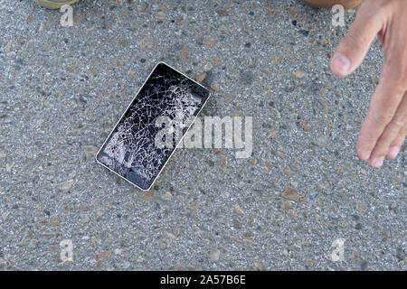 Mann sitzt und halten eine gebrochene smart phone mit einem gesprungenen Bildschirm. Kerl ließ sein Handy auf dem Asphalt und zerbrach es. Ein frustrierter Mann mit Stockfoto