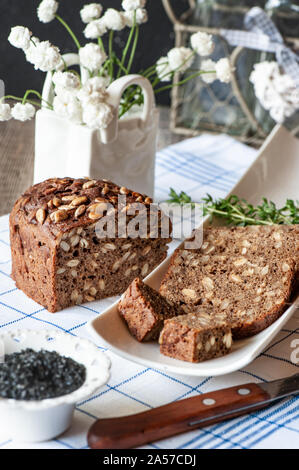 Frisch gebackenem Roggenbrot mit Samen und Rosinen auf sauerteig ohne Hefe auf eine weiße Platte mit einem Messer zum Schneiden und schwarzem Salz. Hausgemachte Kuchen. Stockfoto