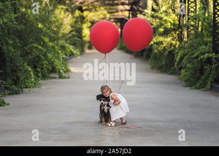 Adorable 4-jährige Mädchen umarmt Hund und halten rote Luftballons Stockfoto