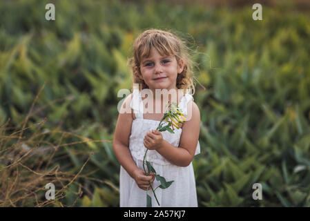 Junge Mädchen in weißem Kleid holding Weinstock mit gelben Blumen im Feld Stockfoto