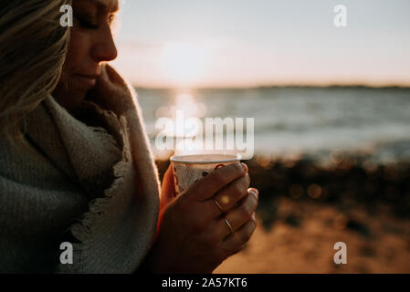 Frau mit Tasse Kaffee am Strand bei Sonnenuntergang Stockfoto