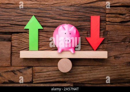 Pfeile nach oben und nach unten und Sparschwein auf Wippe auf hölzernen Schreibtisch Stockfoto