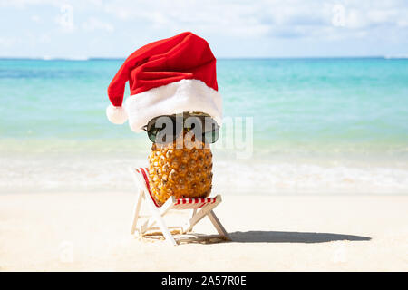 Ananas mit Santa's Hut und Sonnenbrille auf Liegestuhl am Strand in der Nähe des Meeres Stockfoto