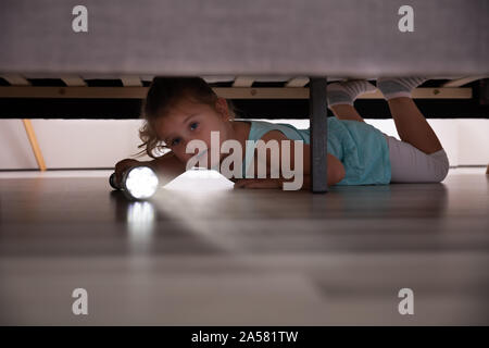 Kleines Mädchen liegt unter dem Bett auf der Suche nach Etwas mit Blitzlicht zu Hause Stockfoto