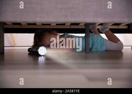 Kleines Mädchen liegt unter dem Bett auf der Suche nach Etwas mit Blitzlicht zu Hause Stockfoto