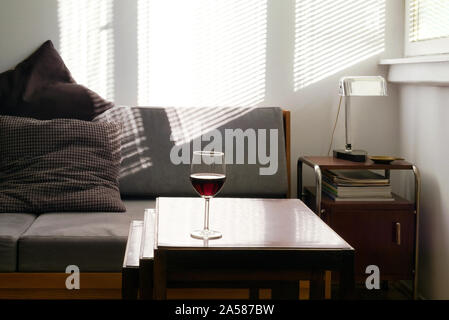 Home Interior am Nachmittag, Sonnenlicht, mit einem Glas Rotwein auf dem Tisch, graue Sofa und Lichtstrahlen auf die Wand im Hintergrund Stockfoto