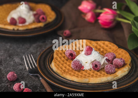 Selbstgemachte Waffeln mit Schlagsahne und organischen Himbeeren. Die Waffeln sind auf zwei braunen Platten auf einem dunkelgrauen Hintergrund, und es gibt einige rote Tul Stockfoto