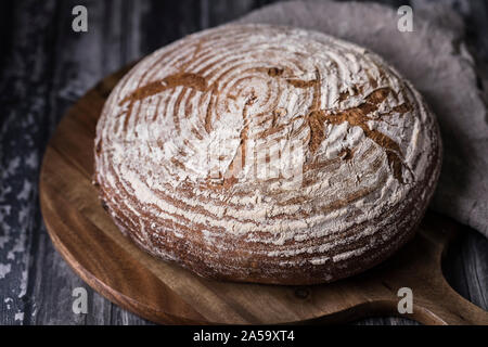 Ein hausgemachtes Brot mit Sauerteig levain Roggen- und Weizenmehl. Das Brot ist auf einem runden Schneidbrett auf einem dunklen Holztisch. Es ist eine braune weiche Bettwäsche Stockfoto