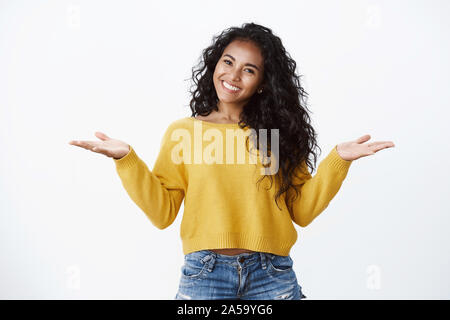 Fröhliche unbeschwerte verlockend Afrikanisch-amerikanische Frau mit Lockigem dunklen Frisur in gelb Pullover, breitete die Hände seitlich zuckend unbothered und lächelnd Stockfoto