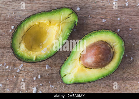 Eine Avocado in zwei mit der Grube in einem der Früchte Hälften aufgeteilt, und einige Meersalz auf dem Holztisch. Die Kamera wird direkt von oben, flach Stockfoto