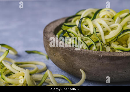 Zucchini Nudeln zoodles in einem rustikalen Schüssel. Die Schüssel ist auf einem blauen strukturierte Oberfläche und es gibt einige geschreddert Zucchini auf dem Tisch neben der Schüssel. Stockfoto