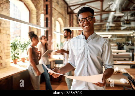Genießen meine Arbeit. Junge afro-amerikanische Mann, Dokumente und Kamera mit Lächeln, während im modernen Büro stehend Stockfoto