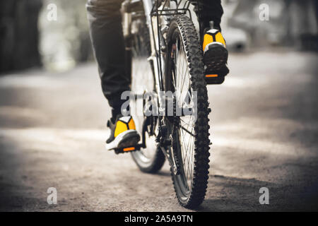 Ein sportlicher Mann in Gelb sneakers reitet ein schwarz Sport Fahrrad mit Pedale mit orangefarbene Reflektoren auf einem sandigen Weg durch Sonnenlicht beleuchtet. Stockfoto